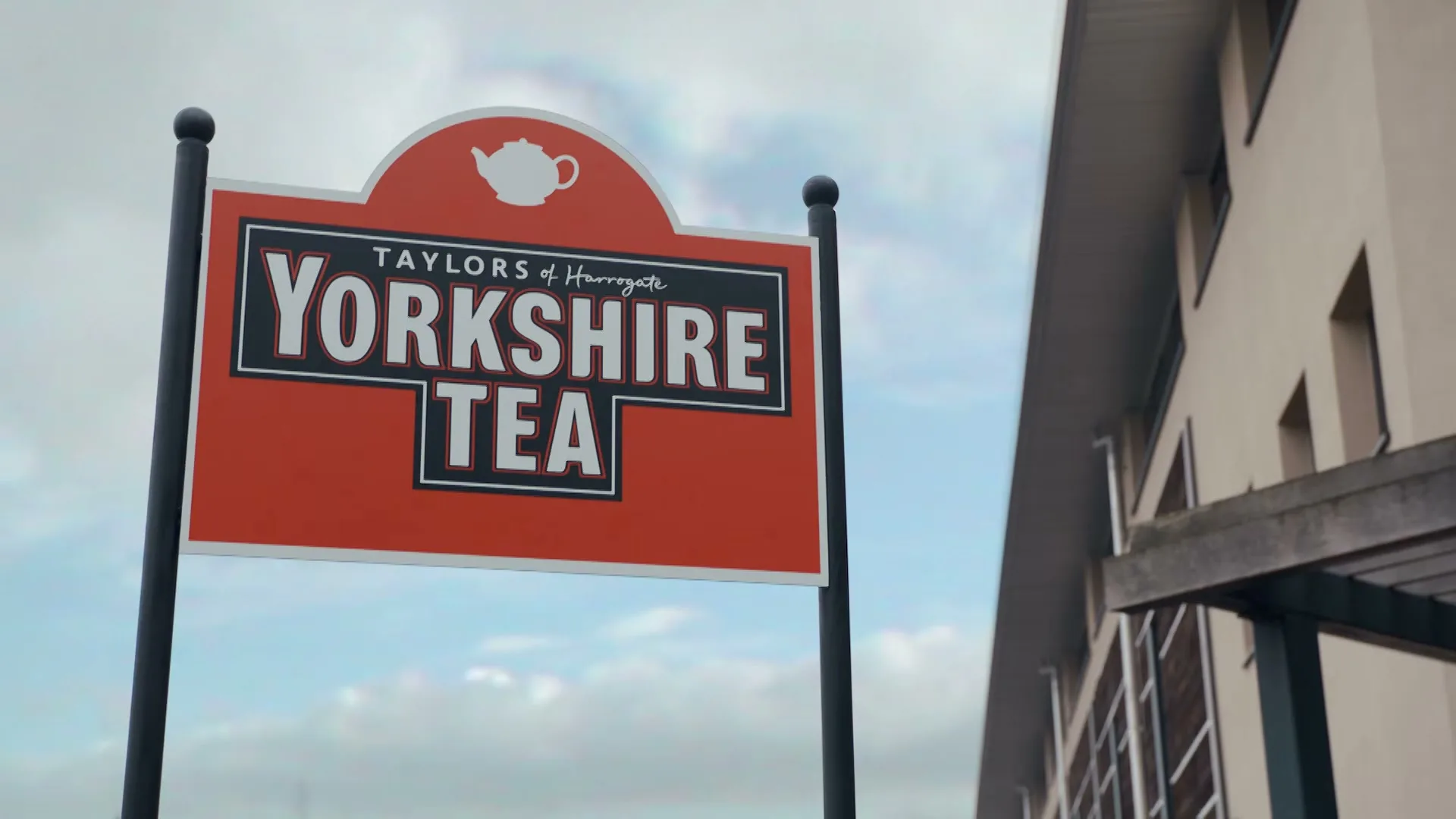 A red and black sign for Taylors of Harrogate Yorkshire Tea stands against a cloudy sky, with a modern building partially visible on the right.