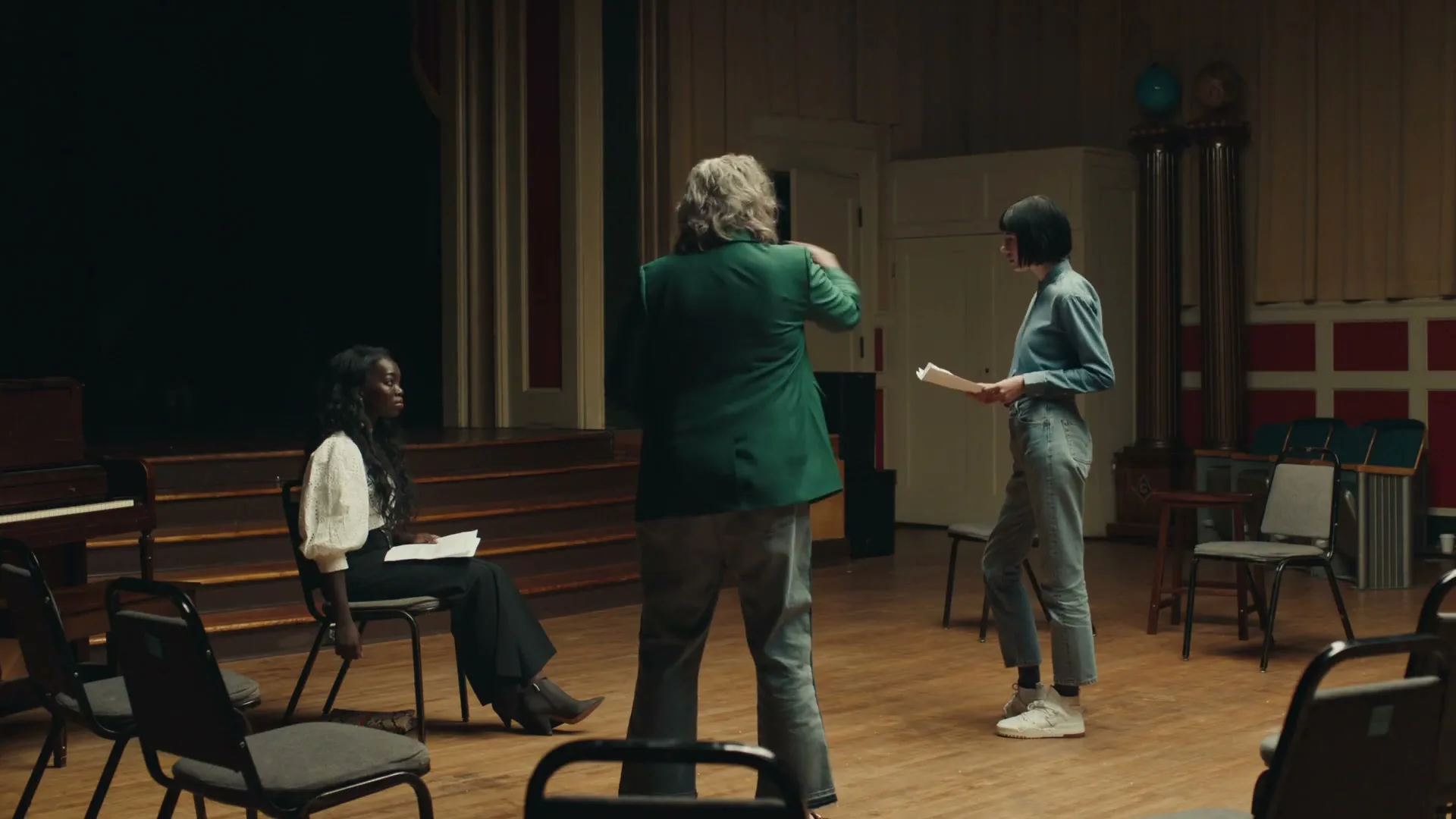 Three people, two women and one man, rehearse a play in a dimly lit auditorium, holding scripts and standing on a wooden floor.