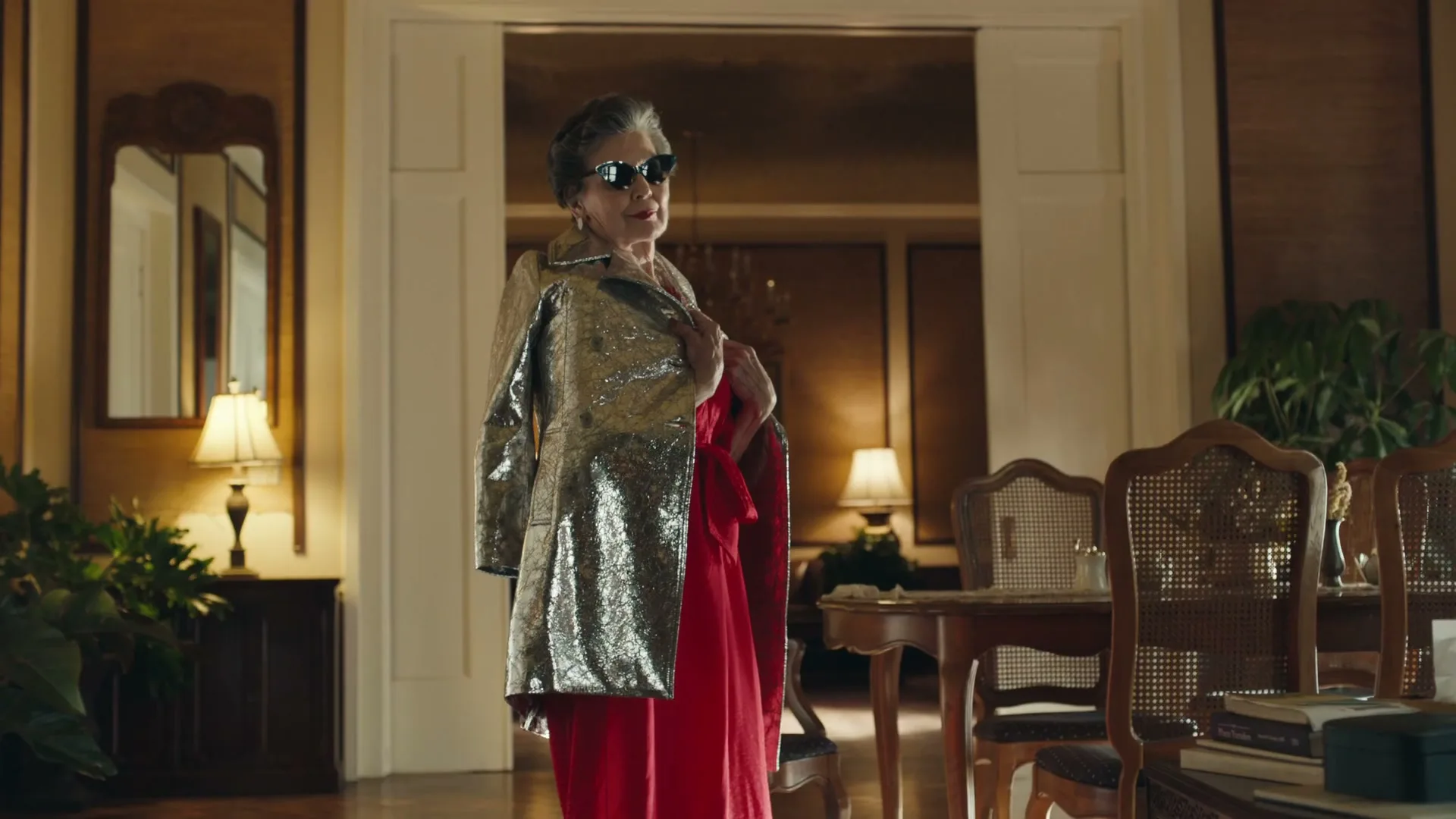 An older woman in a sparkly jacket and red dress poses confidently in a well-decorated room with wooden furniture and lamps.
