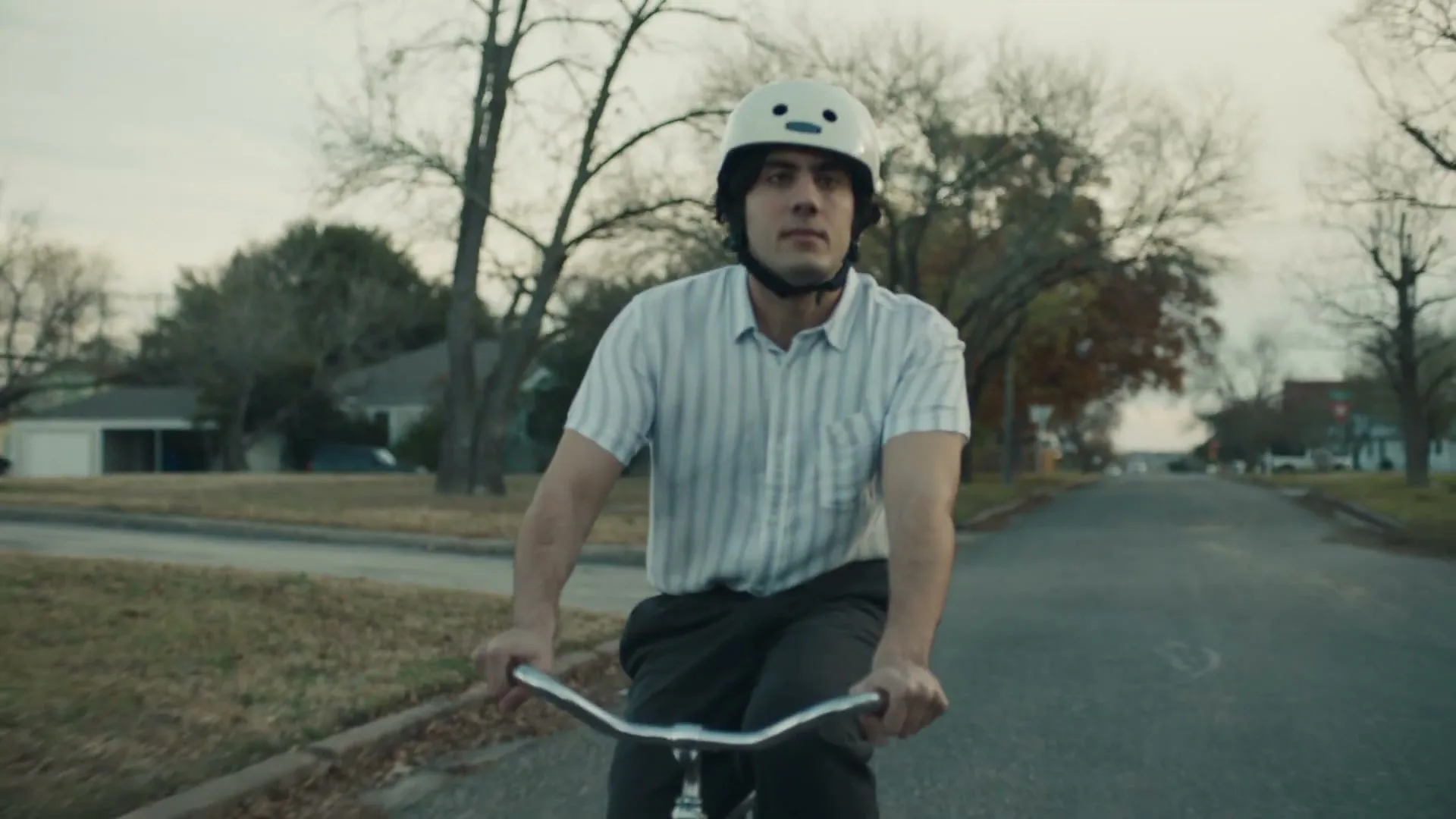 A young man with dark hair and a white helmet rides a bicycle down a suburban street, looking around with a neutral expression.