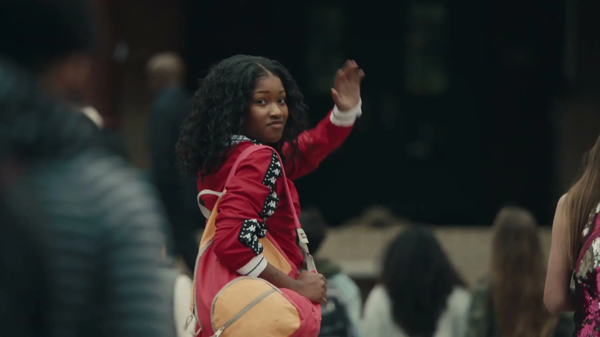 A young Black girl with curly hair, wearing a red jacket and carrying a backpack, waves to the camera in a crowded outdoor setting.