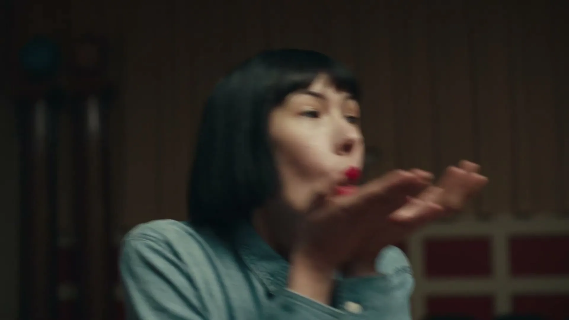 A young woman with short dark hair and red lipstick blows a kiss towards the camera, wearing a denim shirt in a dimly lit room.
