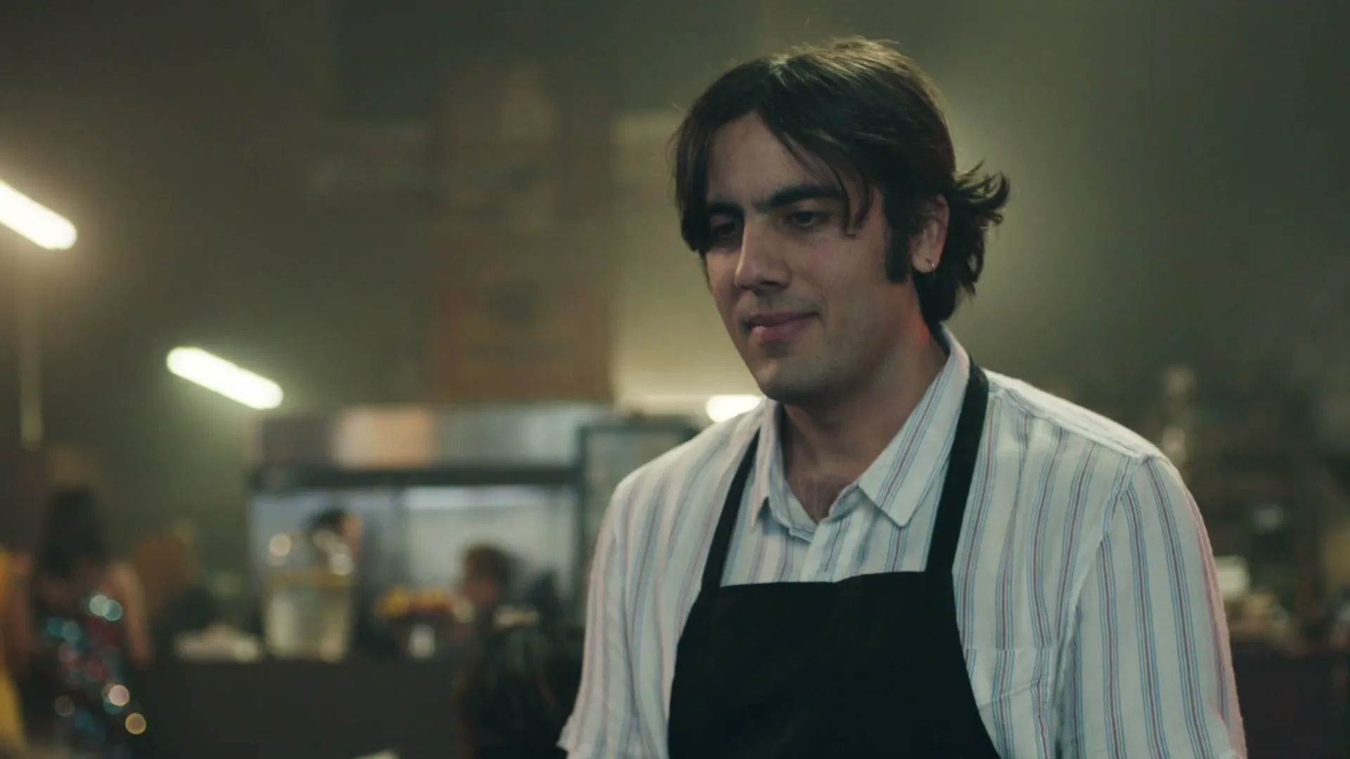 A young male waiter with dark hair and a striped shirt smiles subtly while looking down in a dimly lit restaurant.