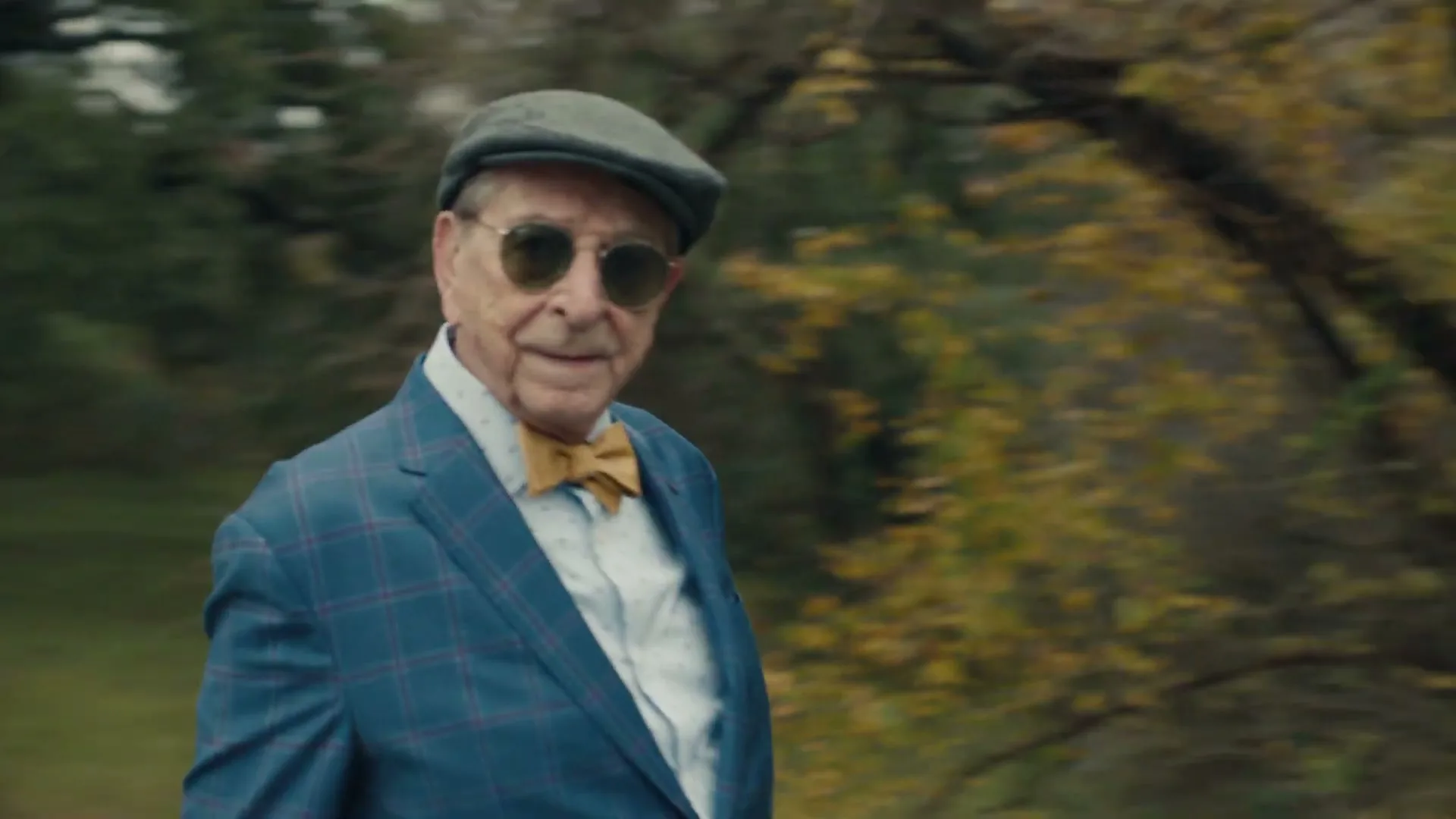 An elderly man in a blue plaid suit, yellow bow tie, and sunglasses walks outdoors, looking at the camera with a slight smile.
