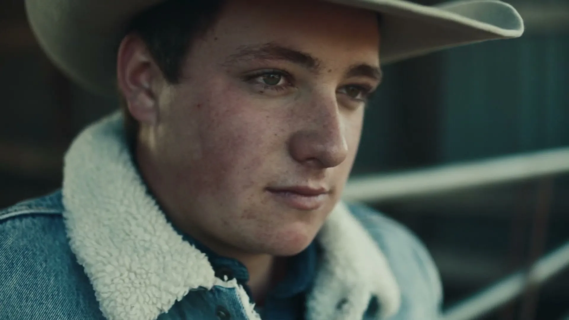 A young male cowboy with a tan hat and denim jacket looks down thoughtfully in an outdoor setting.