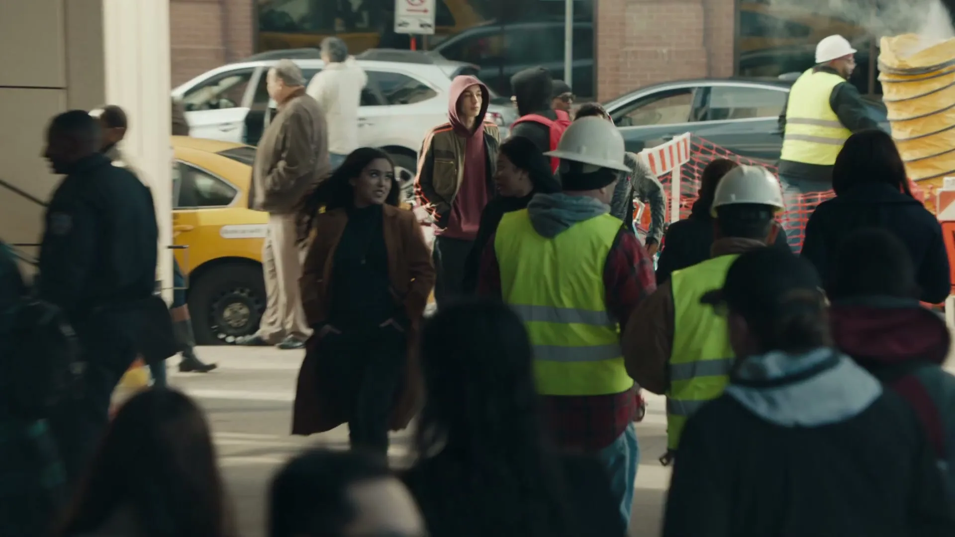 A diverse crowd of people, including construction workers in hard hats and reflective vests, walk through a busy urban area during the day.