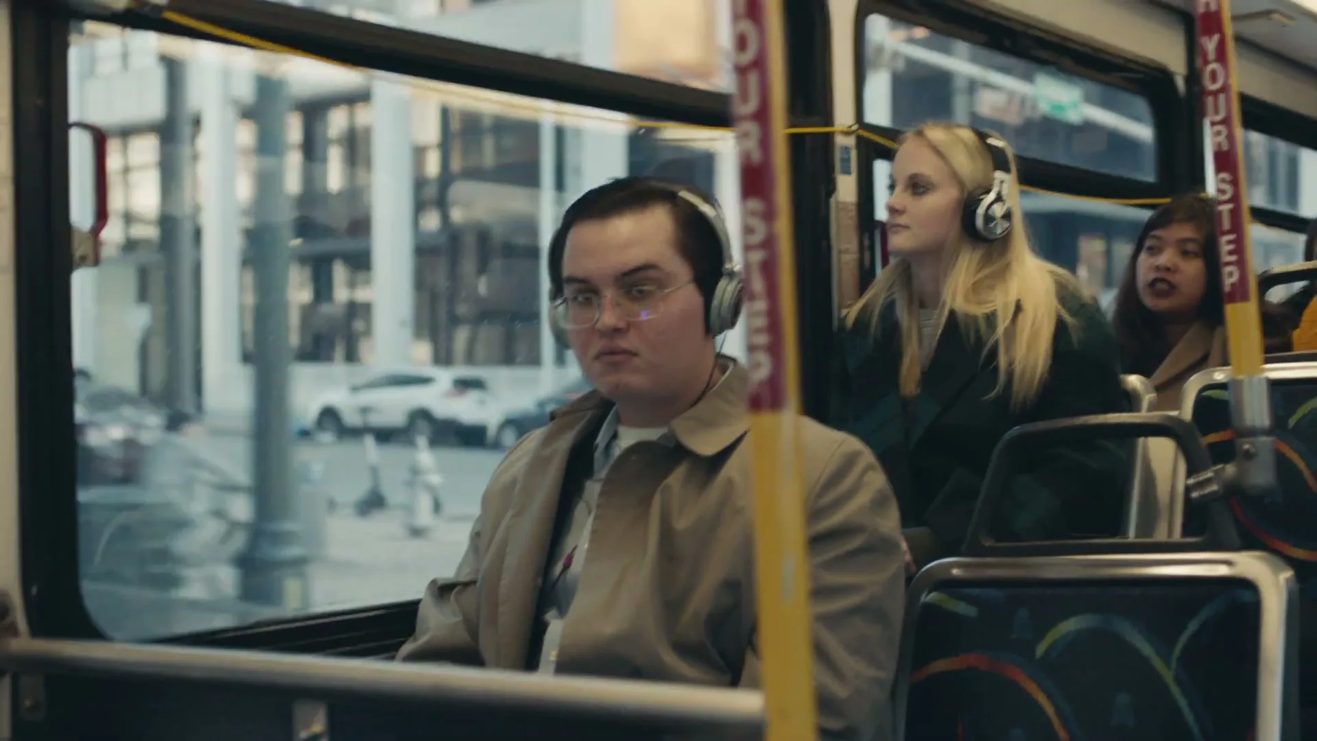 A young man with glasses and headphones sits on a bus, looking out the window, while other passengers are visible in the background.