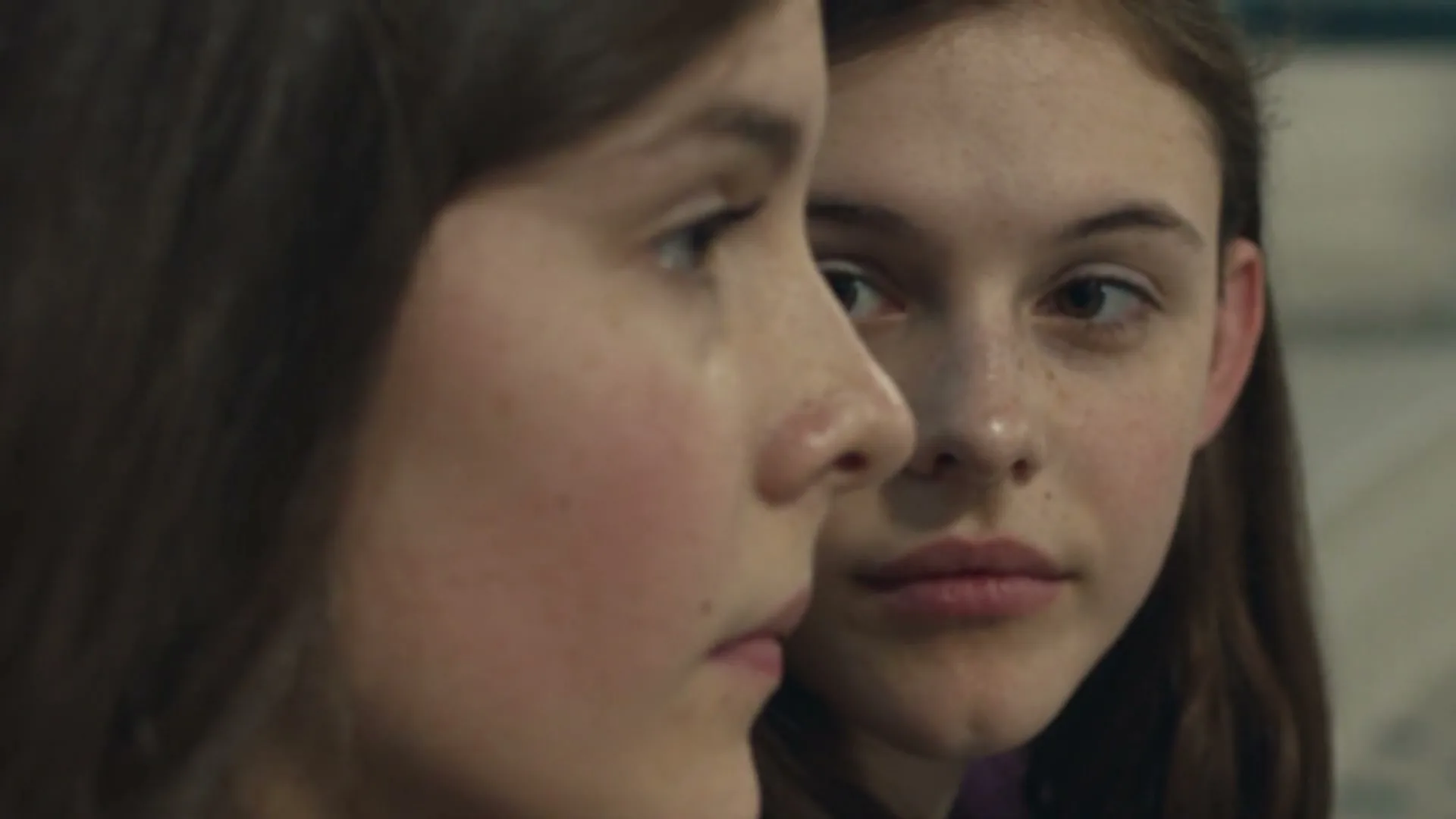Two young women, one in profile and one looking over her shoulder, stand close together in an interior setting.