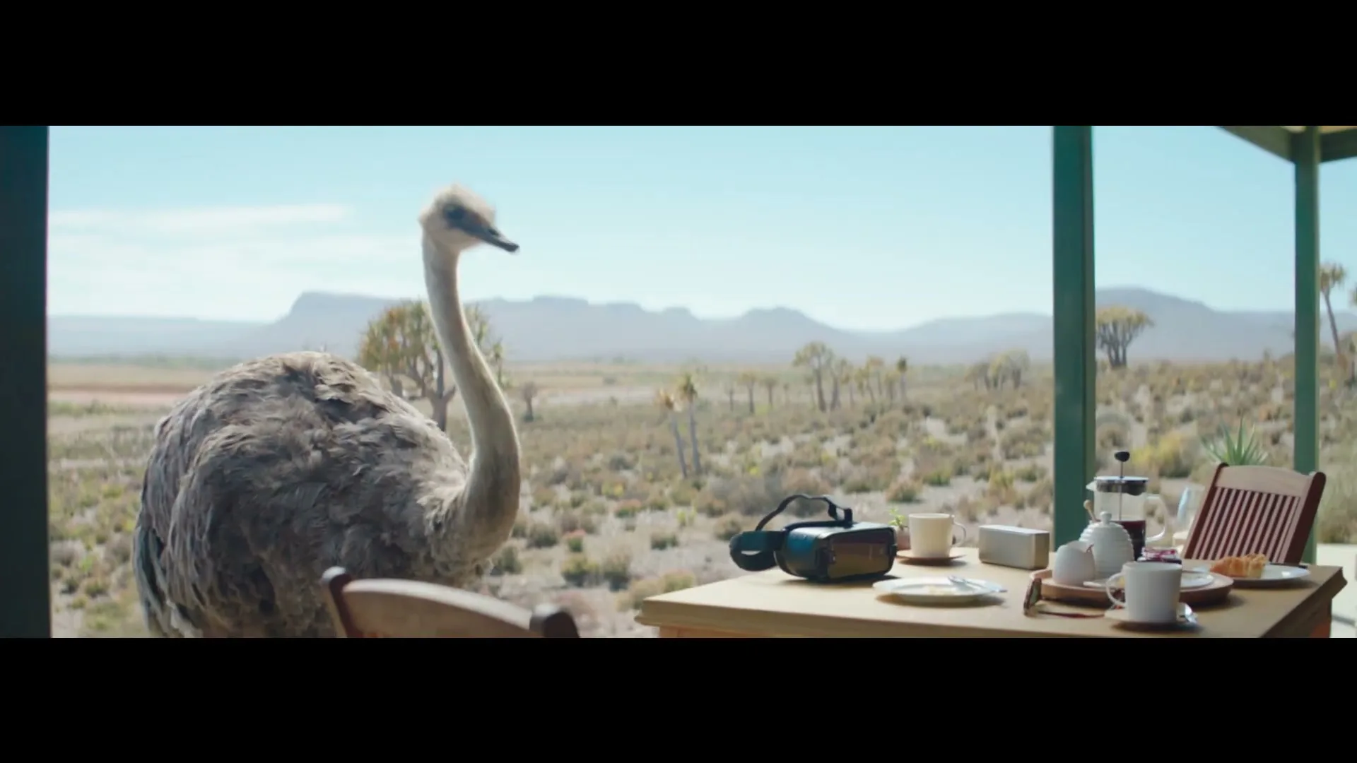 An ostrich looks at a VR headset on a table set for breakfast on a porch overlooking a desert landscape.