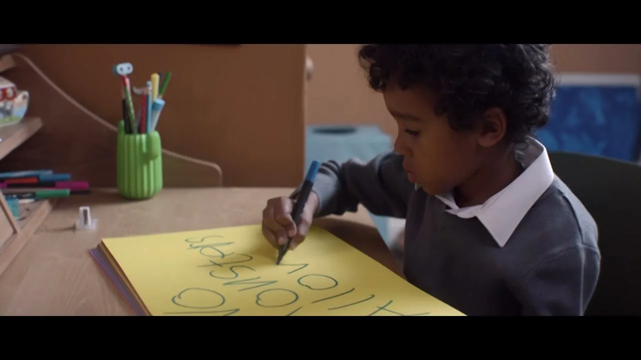 A young boy with curly hair, wearing a grey sweater and white collar, sits at a wooden desk, writing "NO MONSTERS ALLOWED" on a yellow sheet of paper with a blue marker.
