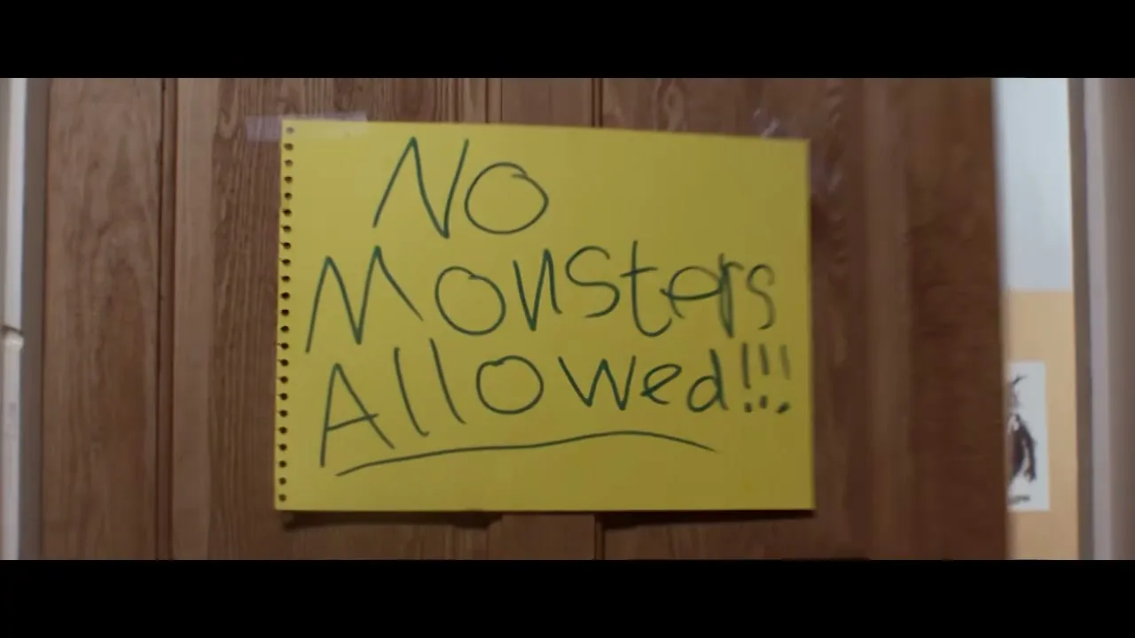 A young boy with curly hair, wearing a grey sweater and white collared shirt, walks past a door with a yellow sign that reads "No Monsters Allowed!!!" in a child's bedroom.