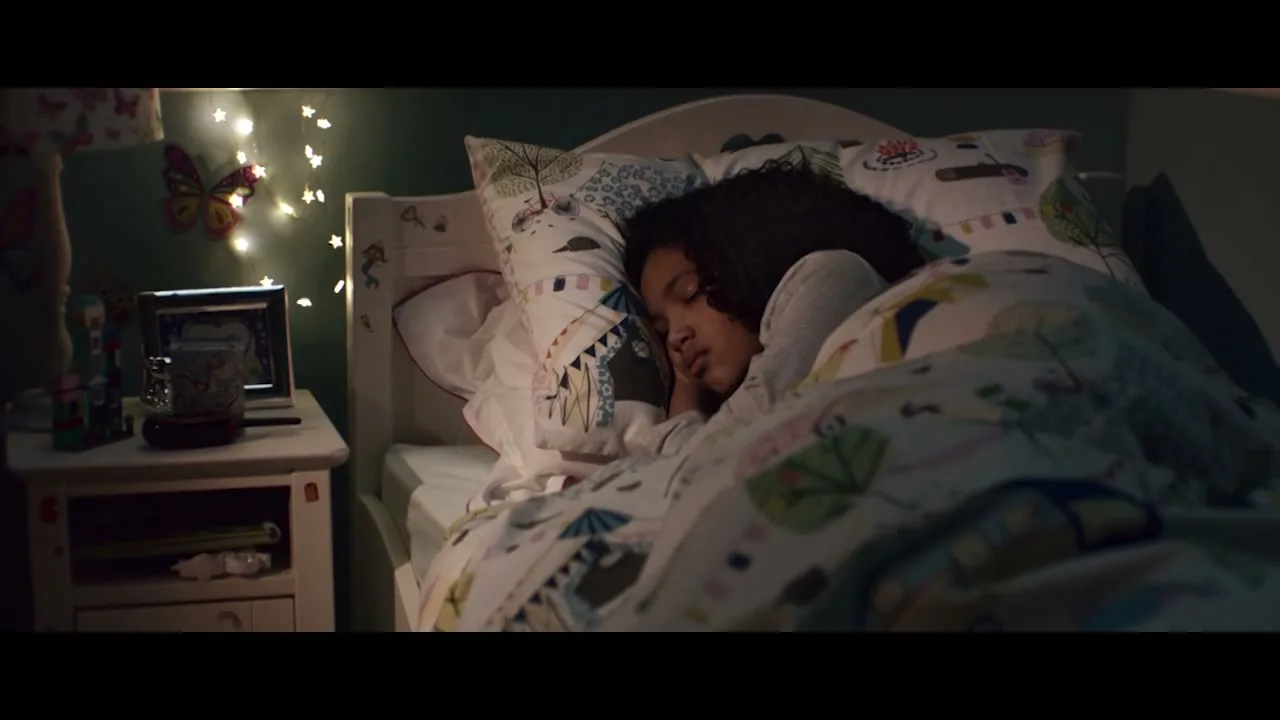 A young girl with dark curly hair sleeps soundly in her bed, covered by a patterned duvet, in a dimly lit bedroom at night.