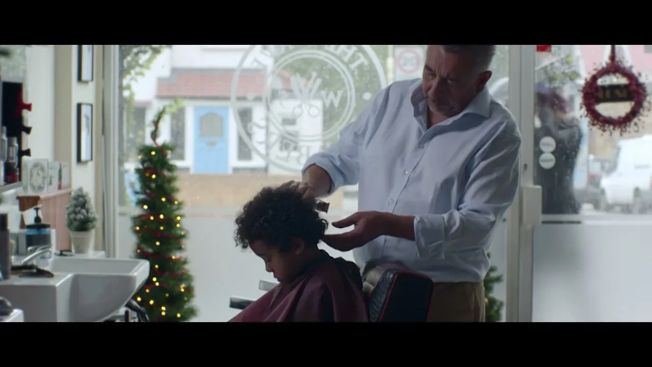 A young boy with curly hair sits in a barber's chair, looking down as a male barber trims his hair in a brightly lit shop decorated for Christmas.