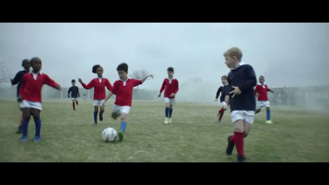 A group of diverse children in red and blue soccer uniforms play soccer on a grassy field under a cloudy sky.