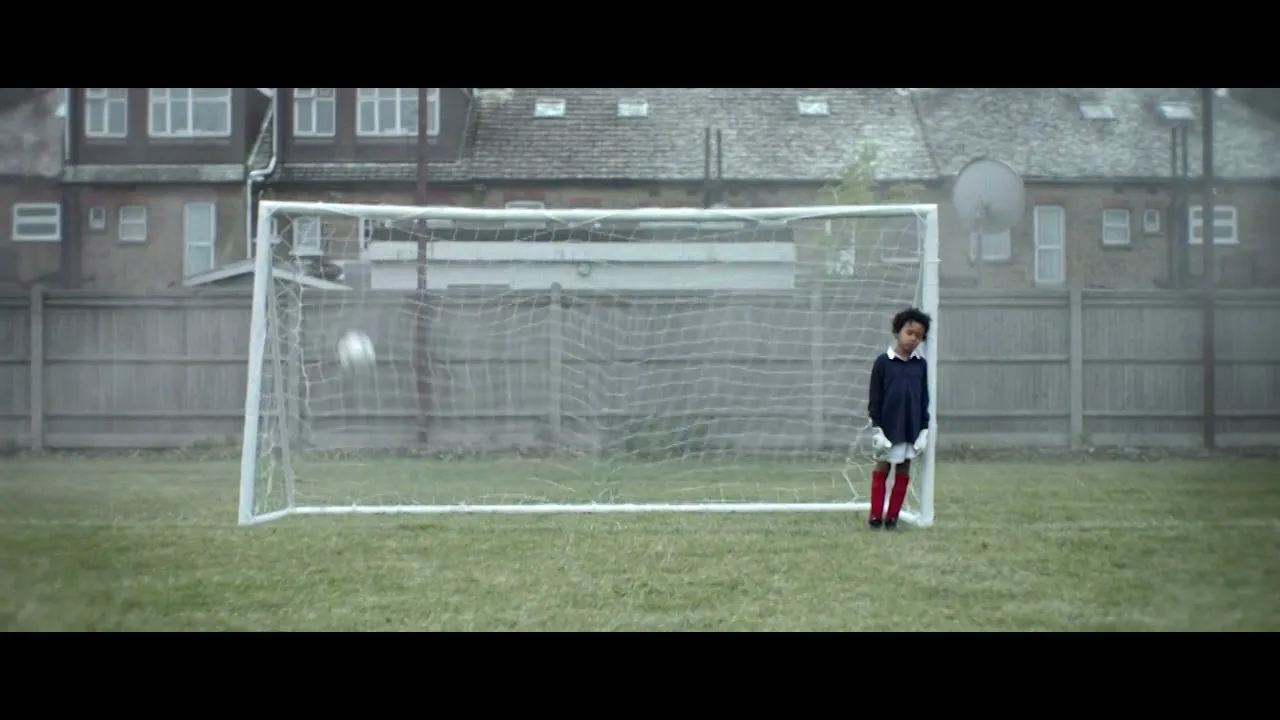 A young boy in a navy shirt and red socks stands next to a soccer goal as a soccer ball flies past him in a grassy field.