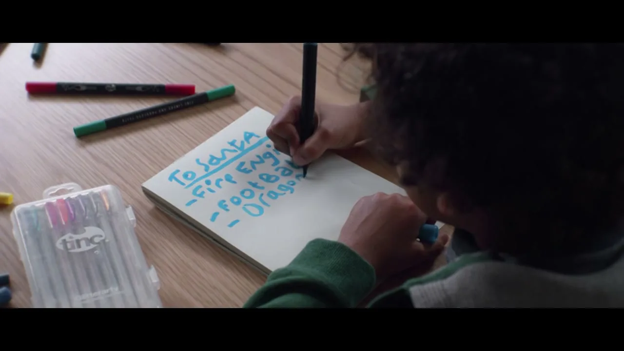 A young boy with curly hair writes a Christmas list to Santa with a blue marker at a wooden table.