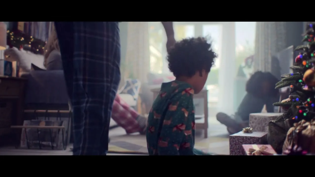 A young boy in pajamas sits on the floor next to a Christmas tree, looking at presents, while a woman and another child are in the background.