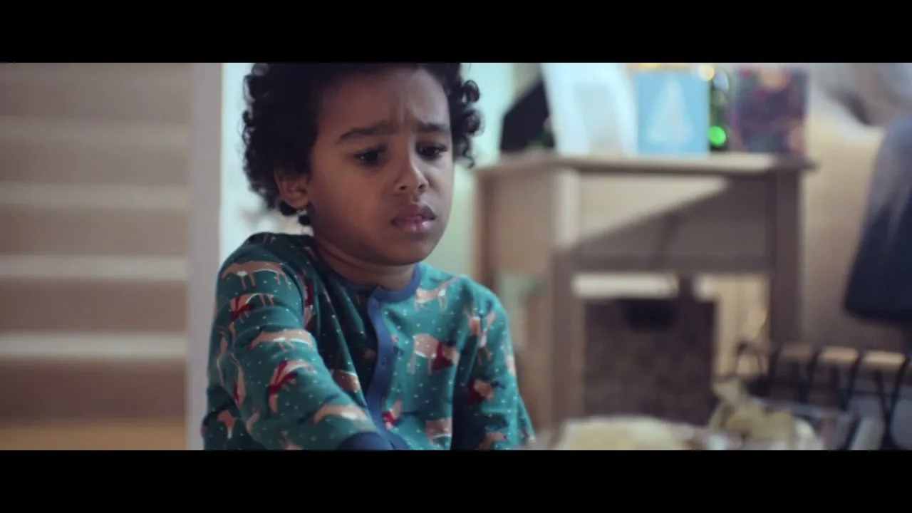 A young boy with curly hair, wearing green pajamas with deer, sits on the floor and unwraps a present in a cozy living room.