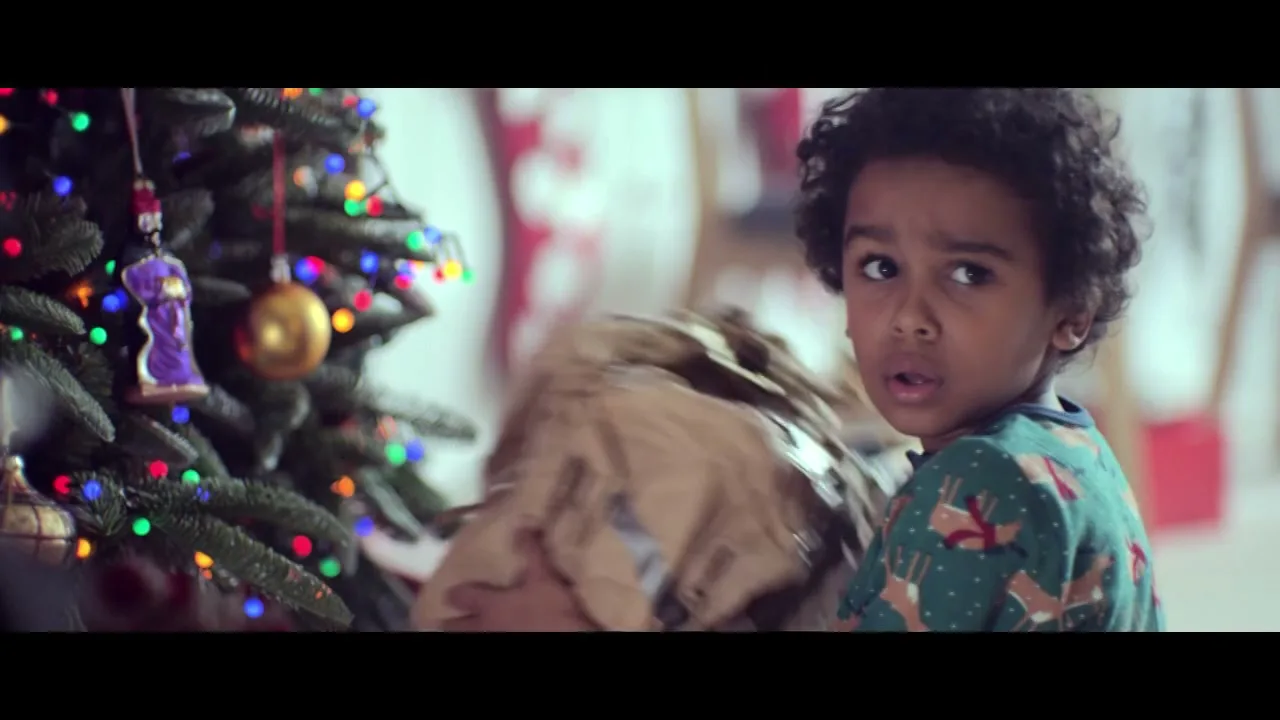 A young boy with curly hair, wearing green pajamas, looks over his shoulder at a Christmas tree in a living room.