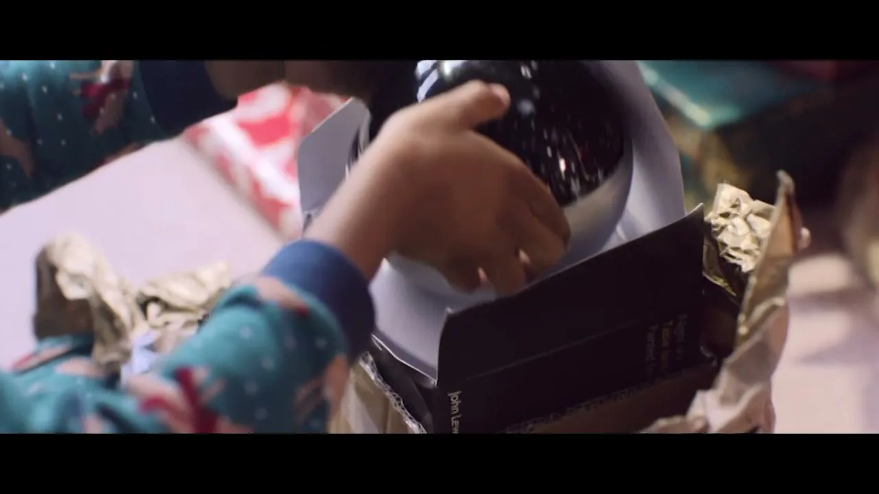 A child's hands in festive pajamas carefully unbox a gift, revealing a star projector inside a cardboard box.