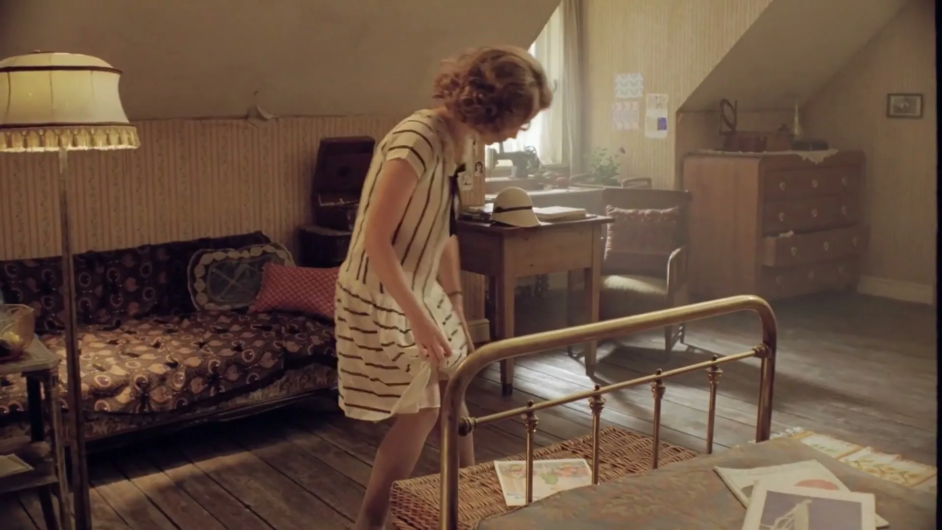 A young woman with curly hair in a striped dress walks into a vintage attic bedroom, bending down to pick something up from the floor.