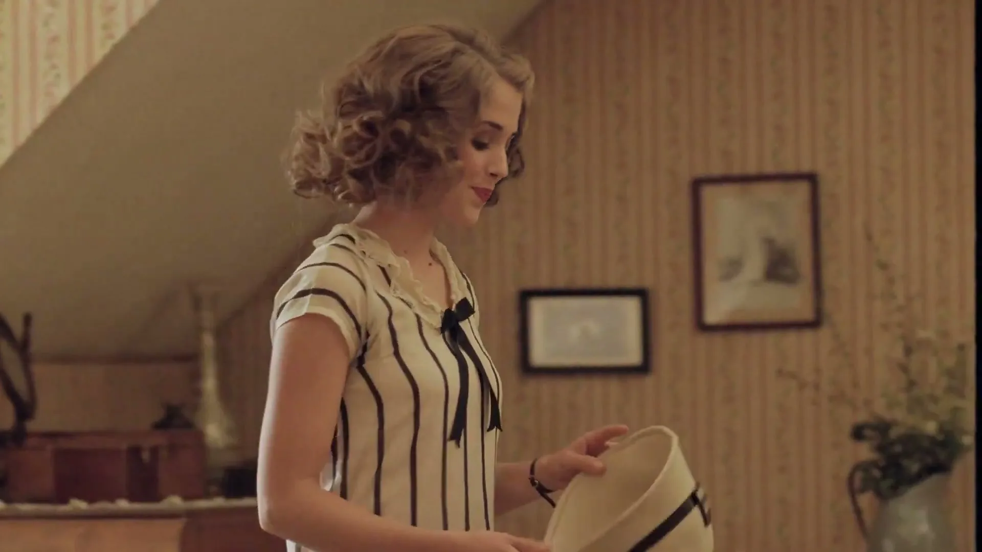 A young woman with curly hair, wearing a striped top, holds a hat and walks away in a room with patterned wallpaper.
