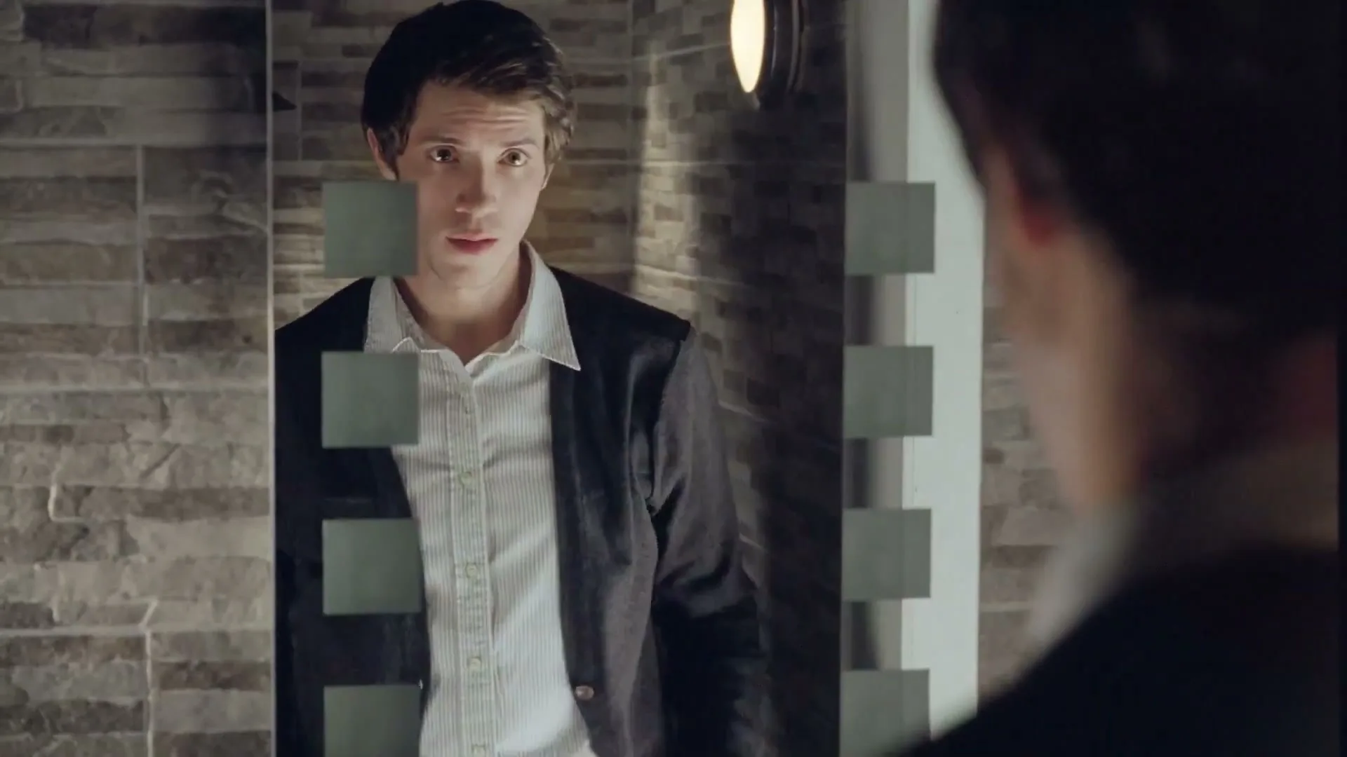 A young man in his 20s, wearing a white shirt and dark cardigan, looks at his reflection in a mirror, adjusting his clothes in a bathroom.