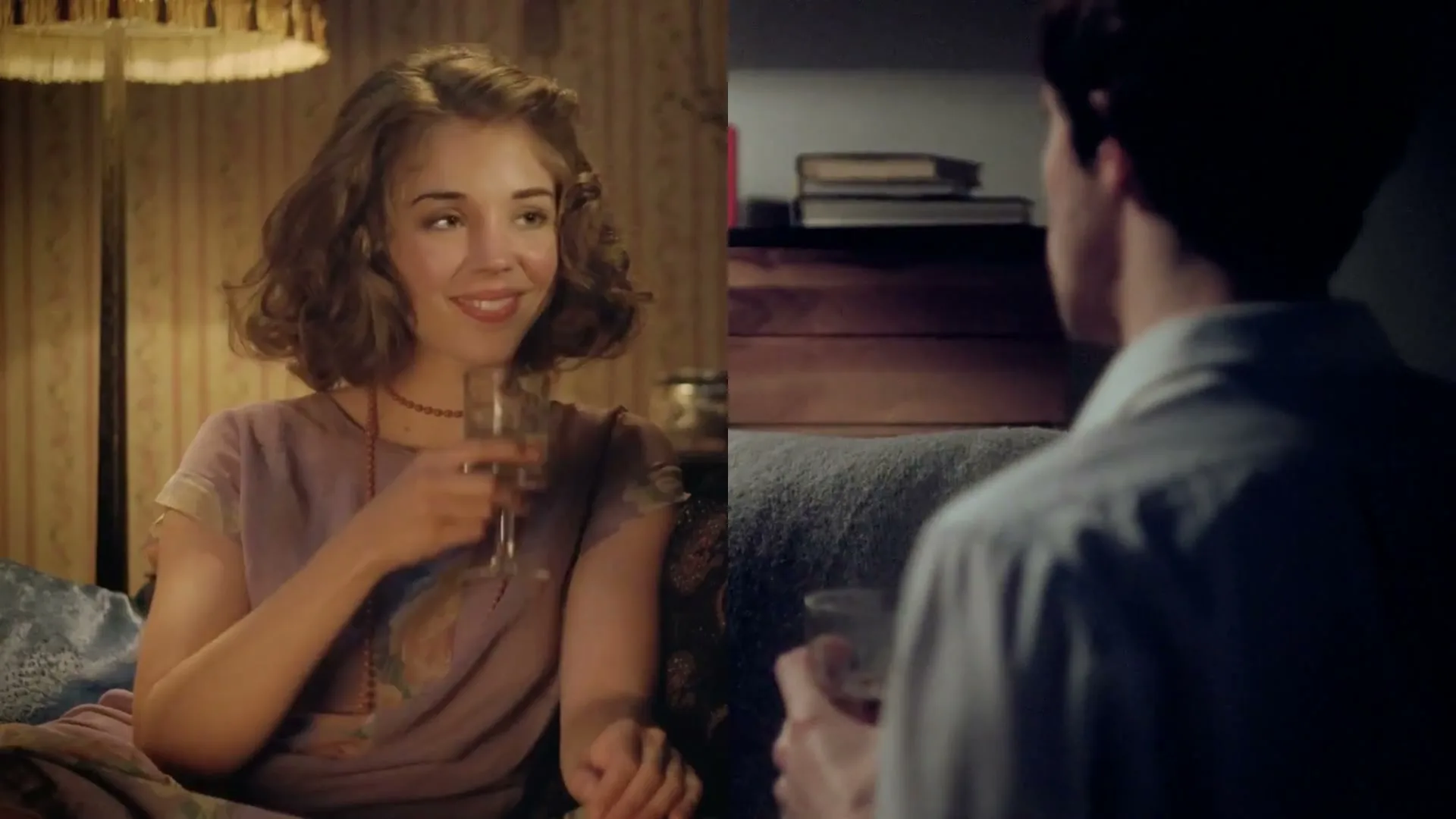 A young woman with curly hair smiles while holding a drink, sitting next to a man in a dimly lit room.