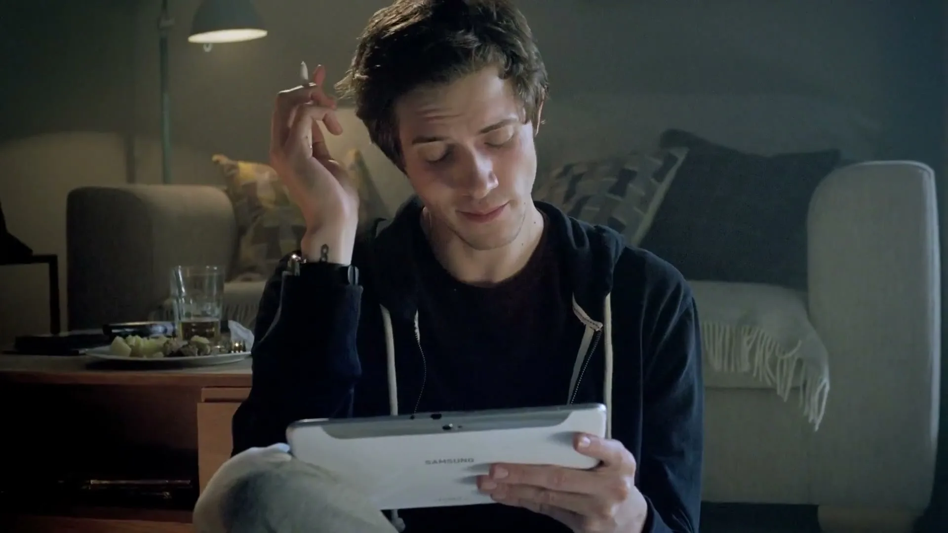 A young man sits on the floor, looking at a Samsung tablet, scratching his head, in a dimly lit living room.
