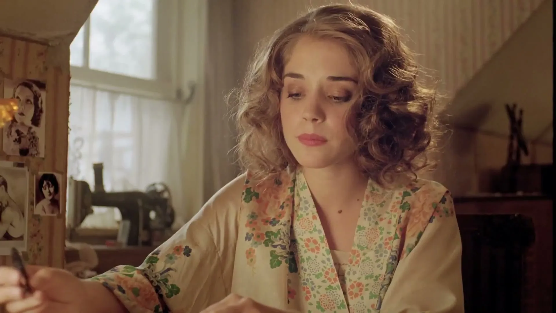 A young woman with curly hair, wearing a floral robe, sits at a desk, looking down and then up, in a sunlit room.