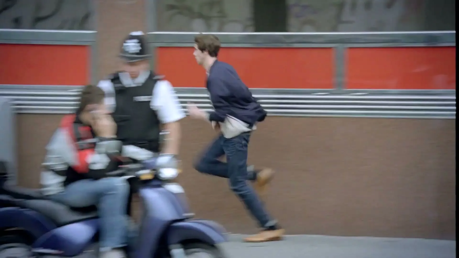A young man in a blue jacket runs past a police officer on a scooter and red recycling bins on a city street.