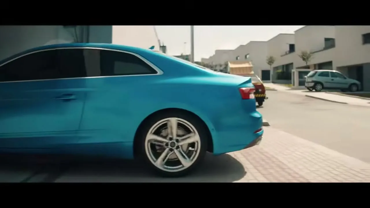 A blue Audi car is parked on a paved area next to a street, with other cars and buildings in the background on a sunny day.