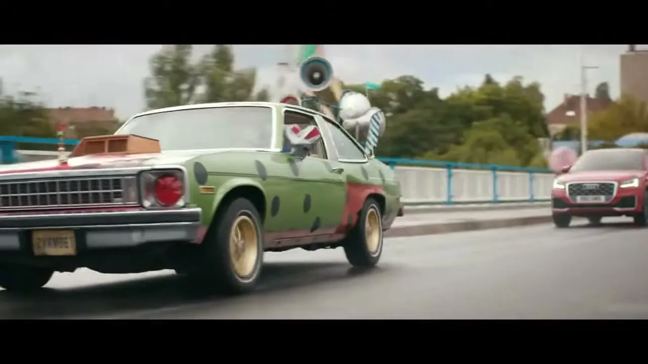 A clown in a green and red polka-dotted car drives on a city street, with a red Audi Q2 driving behind it.