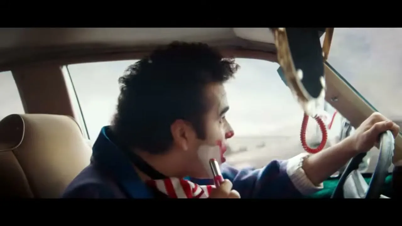 A male clown in a purple jacket and striped tie applies lipstick while driving a car, looking in the side mirror.