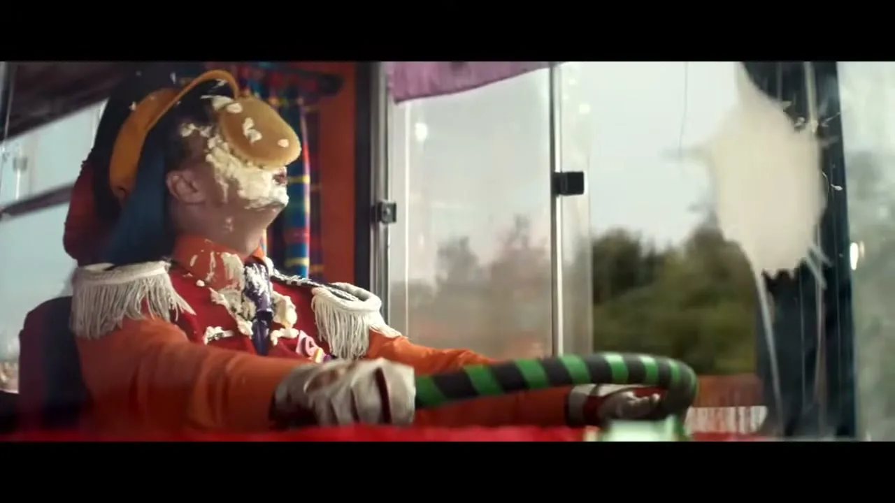 A clown, covered in pie, drives a clown car with a green and black striped steering wheel, looking out the window.