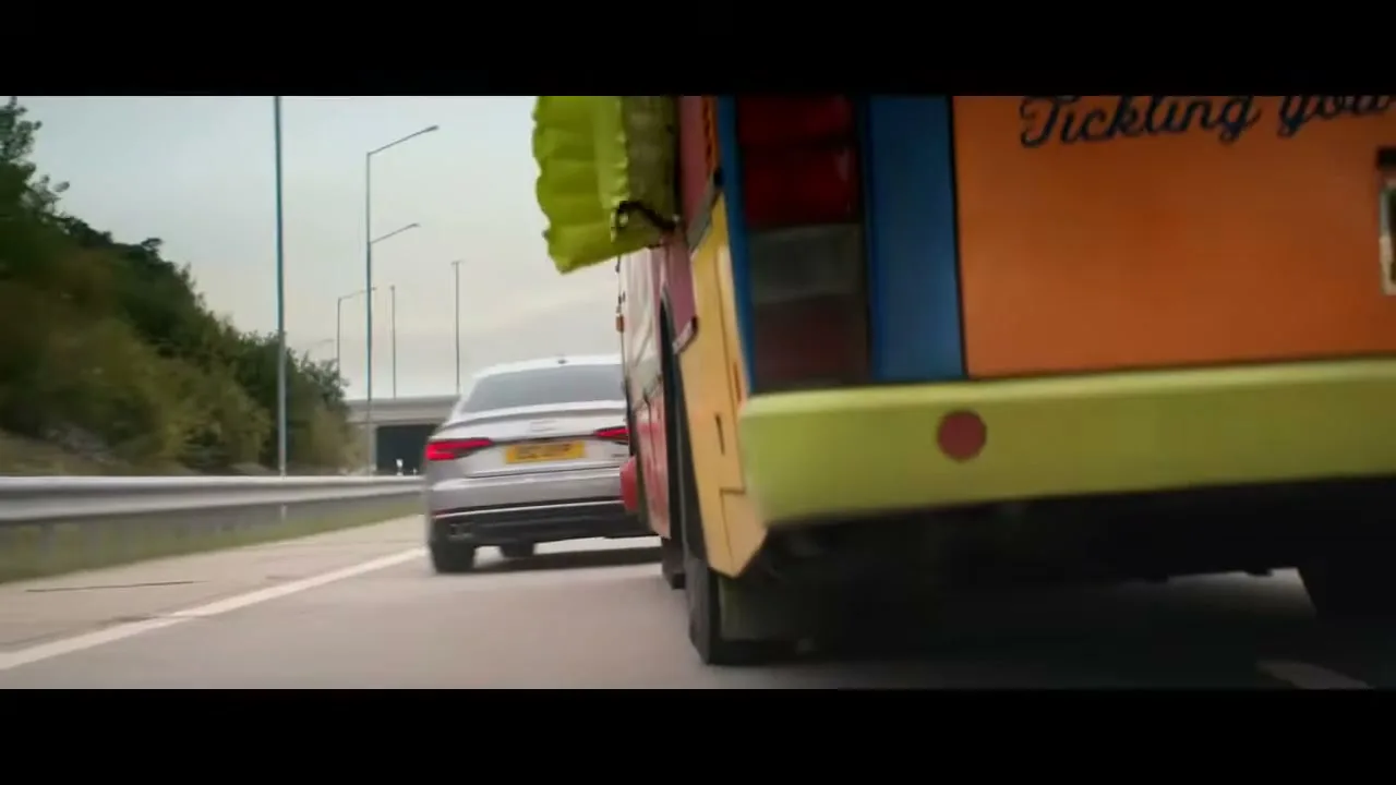 A silver Audi car drives on a highway, passing a colorful clown bus, with an overpass and green hills in the background.