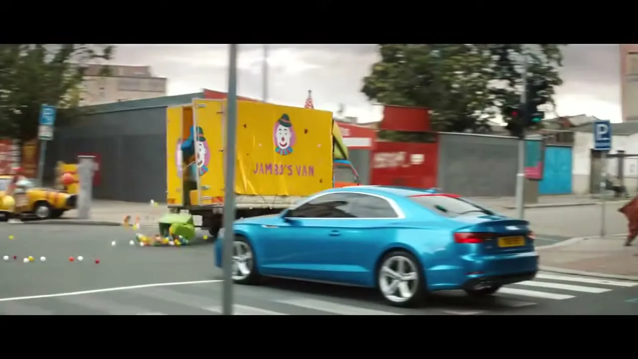 A blue Audi car drives past a yellow clown van that is spilling colorful balls and boxes onto a city street during the day.