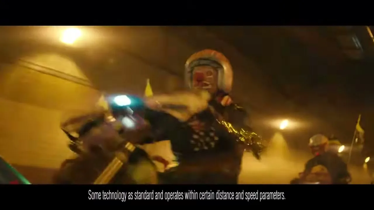 A clown in a helmet and polka-dot jacket rides a motorcycle through a brightly lit tunnel, performing a wheelie.