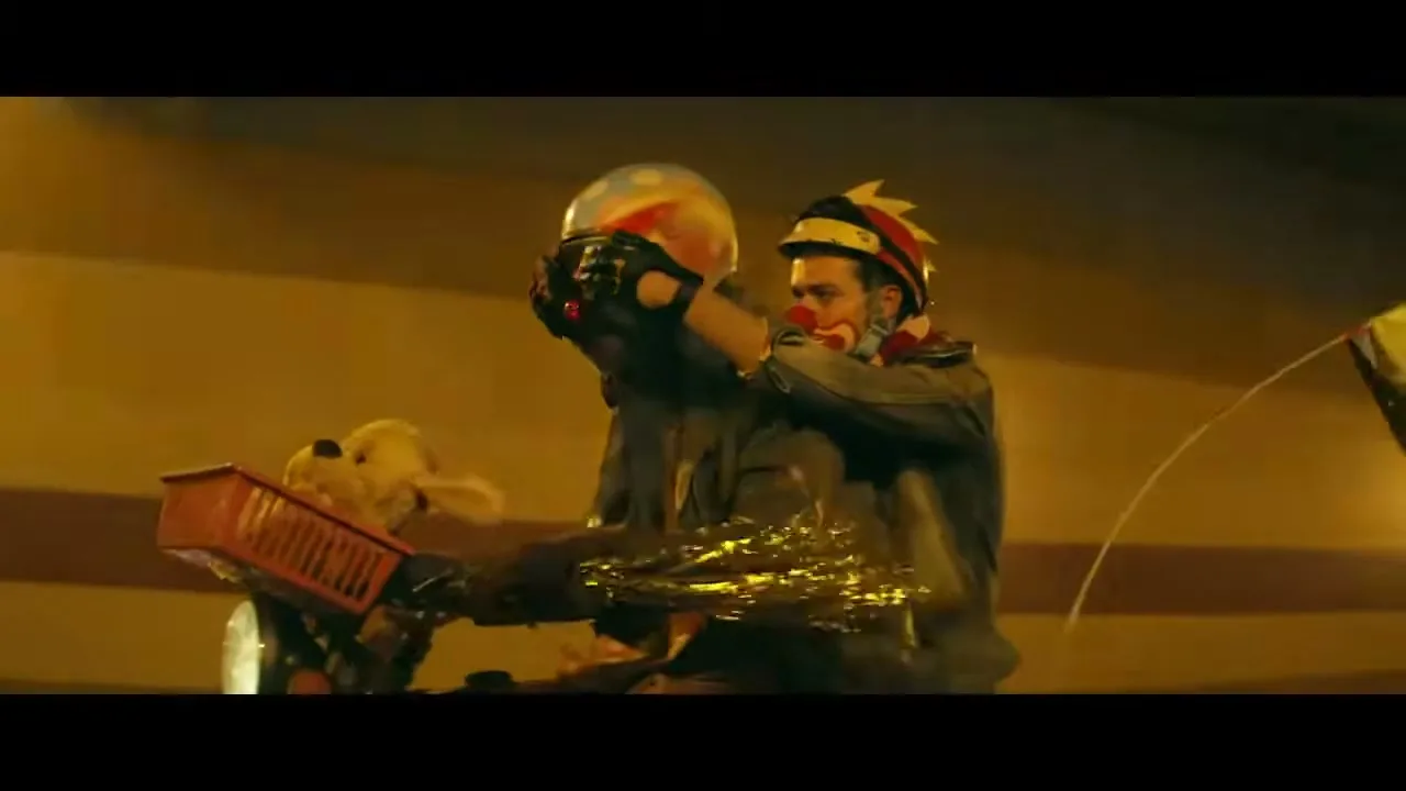 Two clowns on a motorcycle, one wearing a helmet and the other a party hat, ride through a brightly lit tunnel at night.