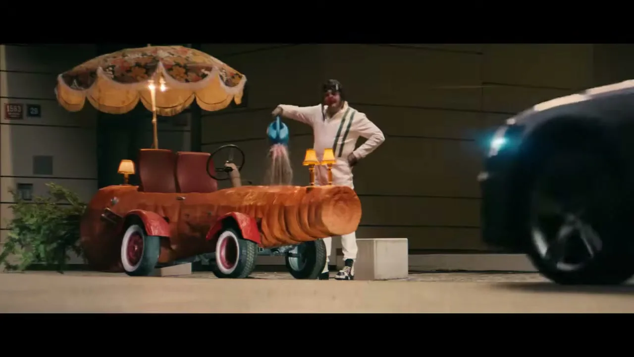 A clown in a white jumpsuit waters a carrot-shaped car with an umbrella and lamps, as a black Audi drives past.