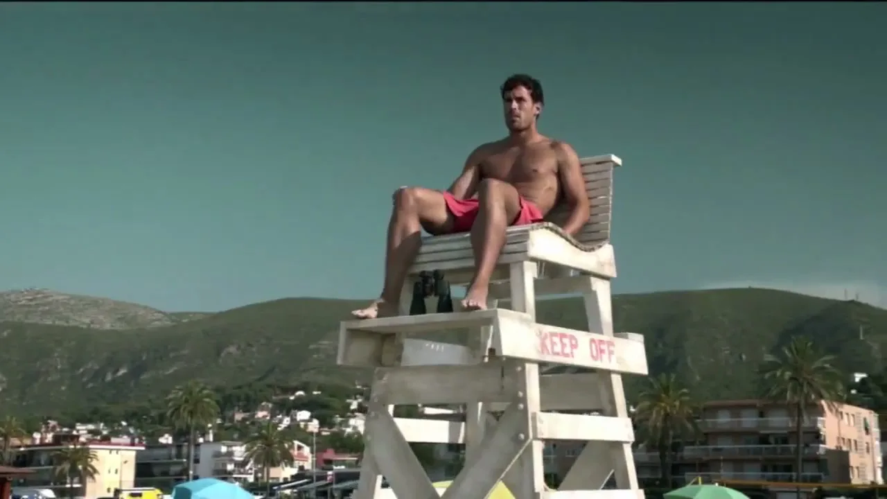 A muscular male lifeguard in red swim trunks sits on a tall wooden lifeguard chair on a beach, looking around.