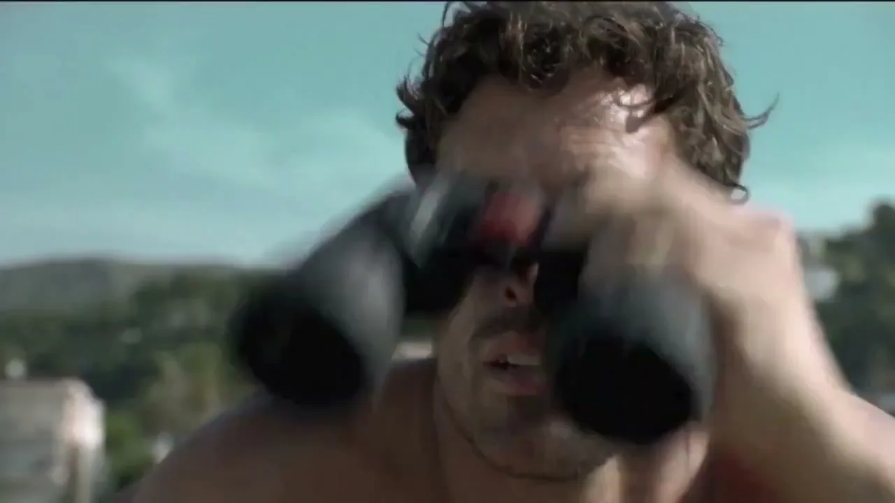 A man with wet, curly hair and a worried expression looks through binoculars, scanning the horizon under a bright sky.