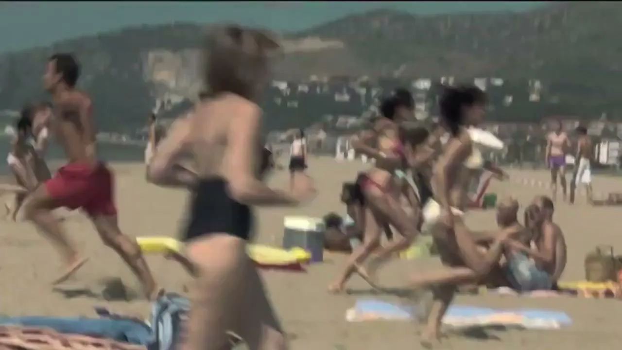A diverse group of young adults in swimwear run towards the ocean on a crowded beach, with mountains in the background.