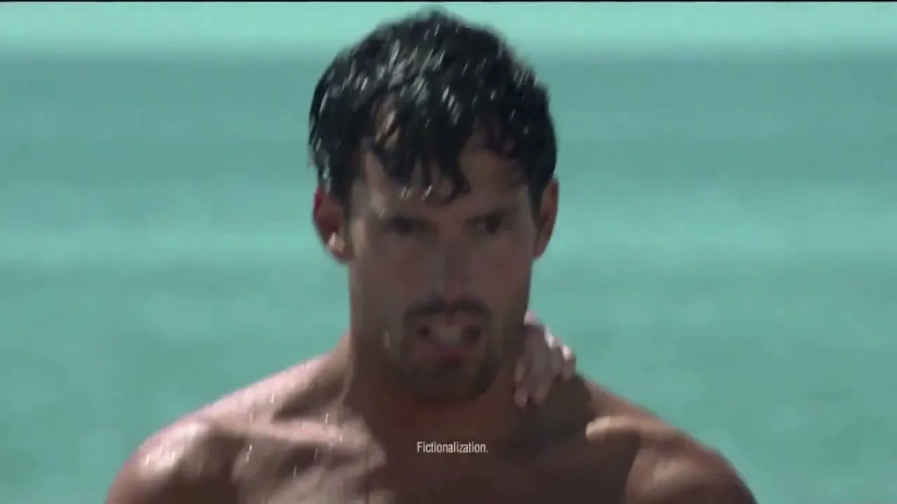 A wet, muscular man with dark hair and stubble looks intensely at the camera while a woman's hand rests on his shoulder in the ocean.