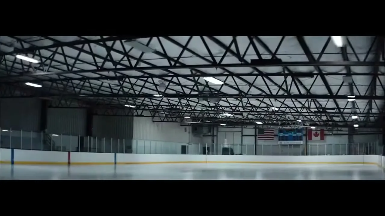 The lights in a dark, empty ice hockey arena slowly turn on, revealing the rink and the metal ceiling structure.