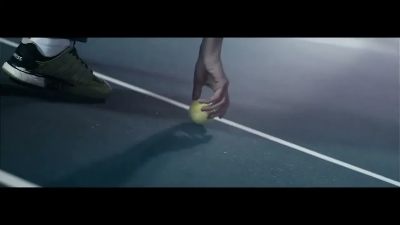 A tennis player's hand reaches down to pick up a tennis ball on a blue court with white lines.