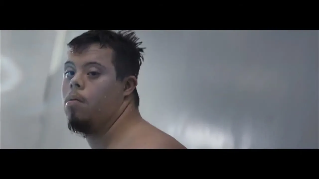 A young man with Down syndrome, shirtless and wet, looks over his shoulder in a shower, with water droplets on his face.