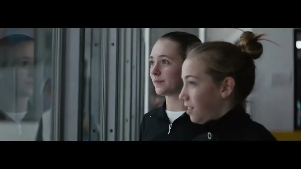 Two young female athletes in black tracksuits look out a window, their reflections visible, in an indoor sports facility.