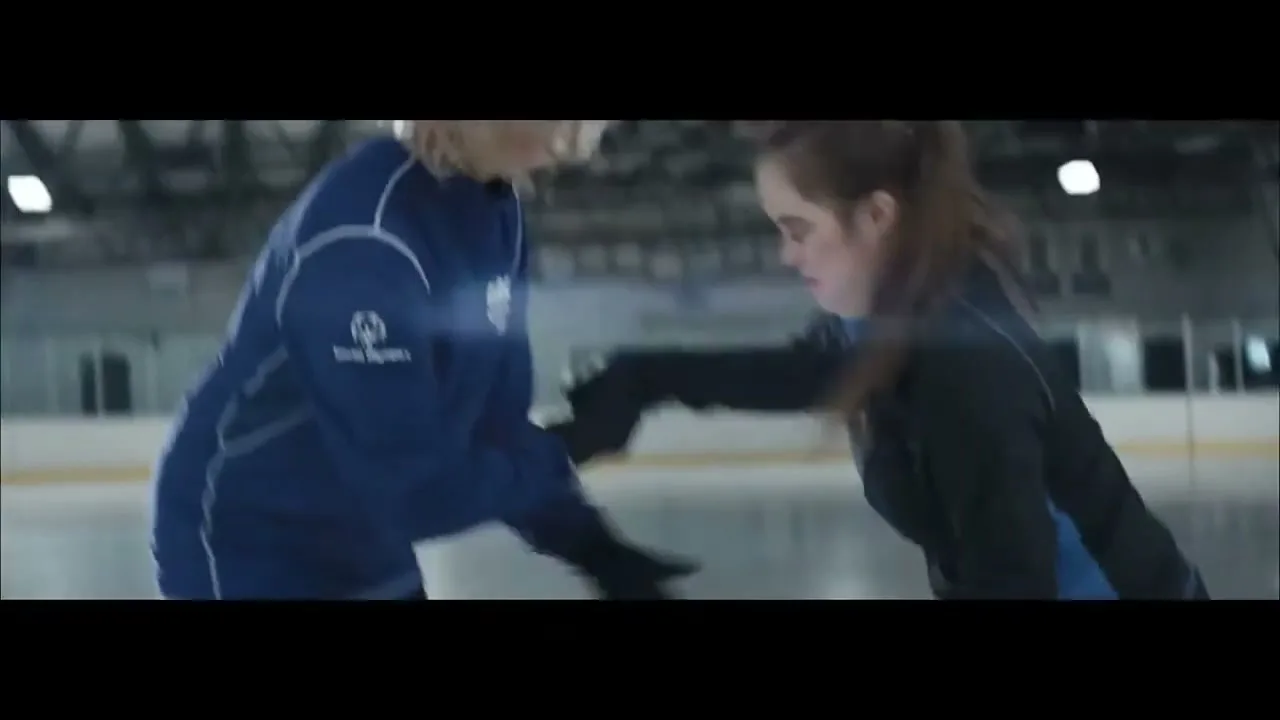A young female ice skater, wearing a black and blue tracksuit, is helped up by an older female coach on an ice rink.