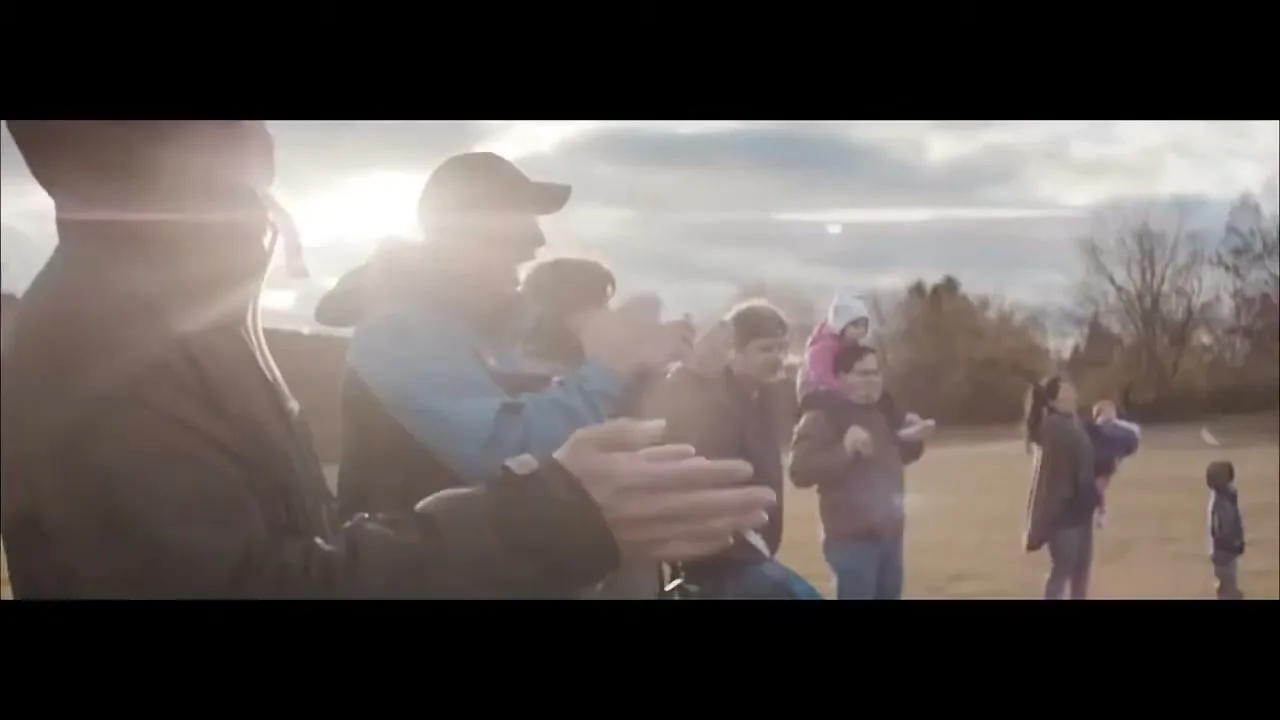 A diverse group of people, including adults and children, stand outdoors on a grassy field, clapping and cheering with joyful expressions under a cloudy sky.